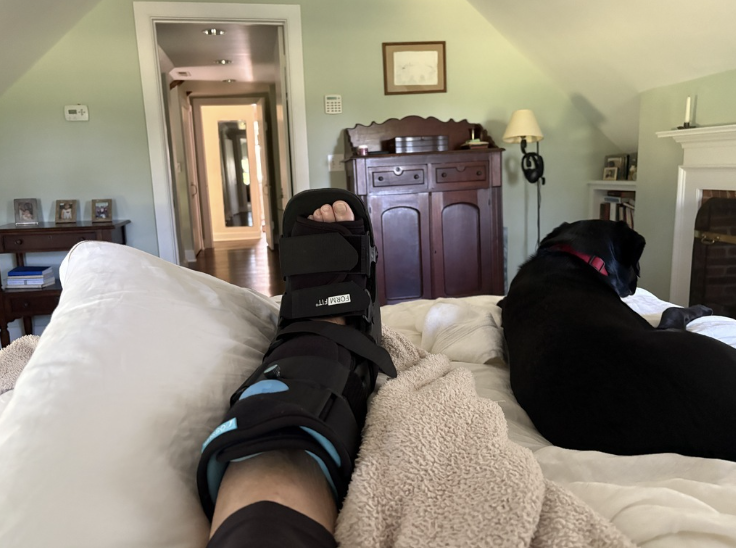 My leg in a black walking boot propped on the bed, with our black lab lying beside me